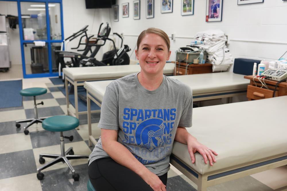 A smiling woman in a "Spartans" t-shirt sits in a physical therapy room. She leans on a treatment table, with more tables and exercise equipment behind her. The room has a blue and white tiled floor.