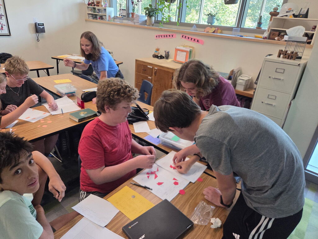 Students gathered around paper cutouts of various shapes to create composite figures. Students are smiling and moving shapes.