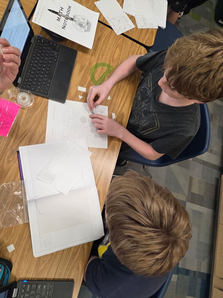Students gathered around paper cutouts of various shapes to create composite figures. A computer is open next to a protractor and student Math notebook.