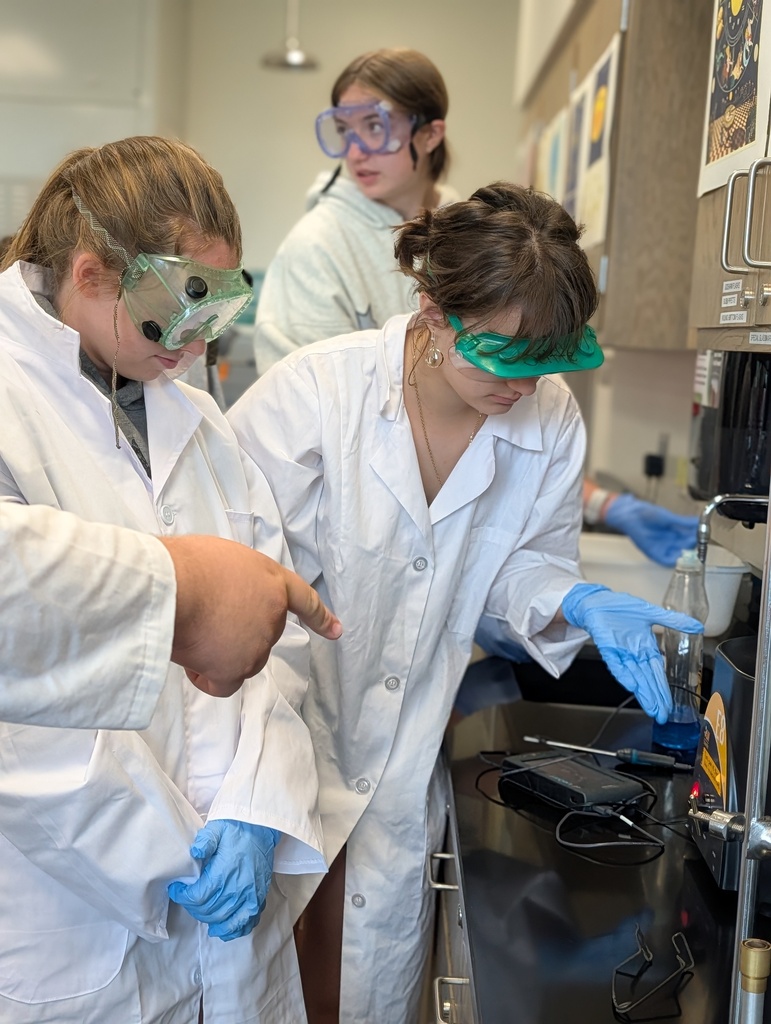 Students wearing goggles, lab coats, and gloves start a centrifuge for a chemistry lab.