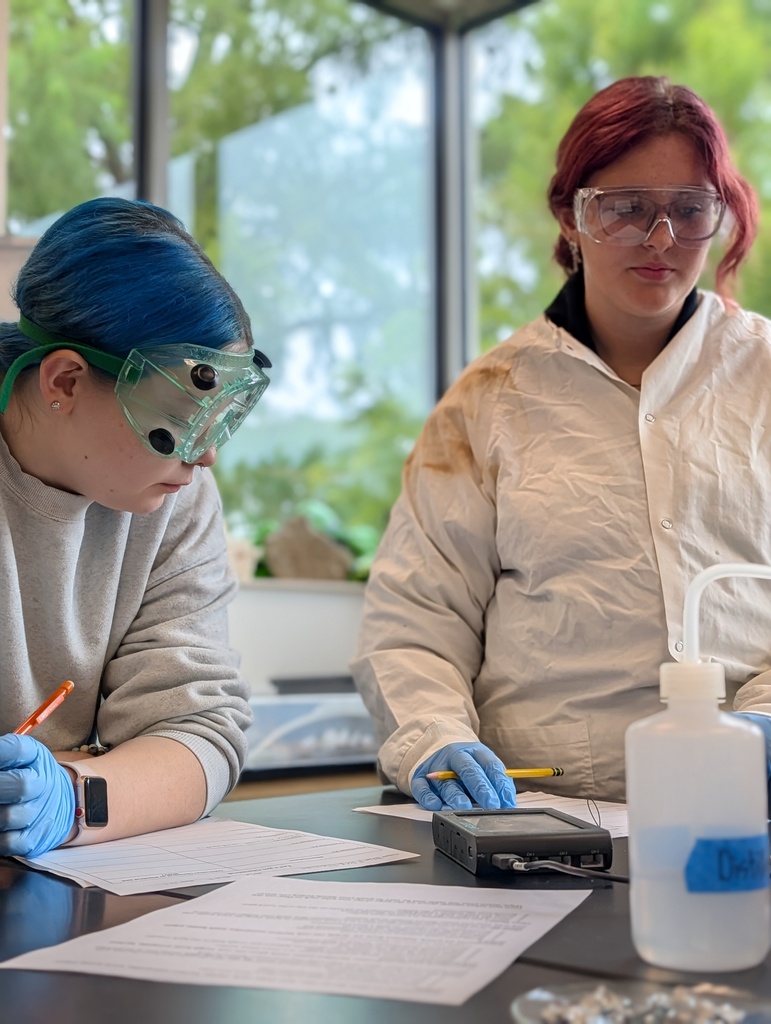 Students wearing lab goggles and blue nitrile gloves conduct a chemistry lab with large glass windows in the background.