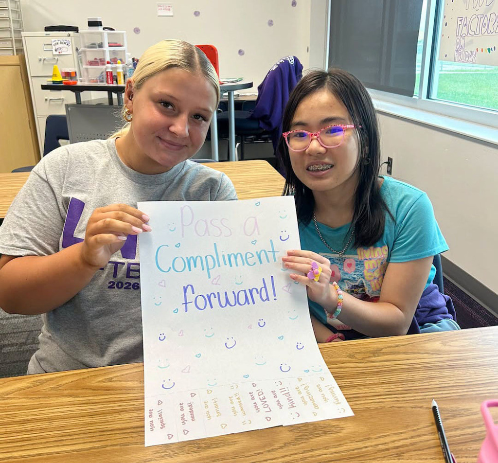 Two Northwest High School students are sitting at a desk in their classroom. The two girls are holding a sign that says, "Pass a Compliment Forward!"