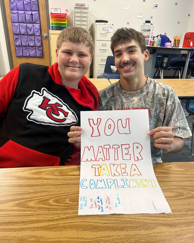 Two Northwest High School students smile as they hold a sign while sitting at a classroom table. The sign says, "You Matter - Take a Compliment."