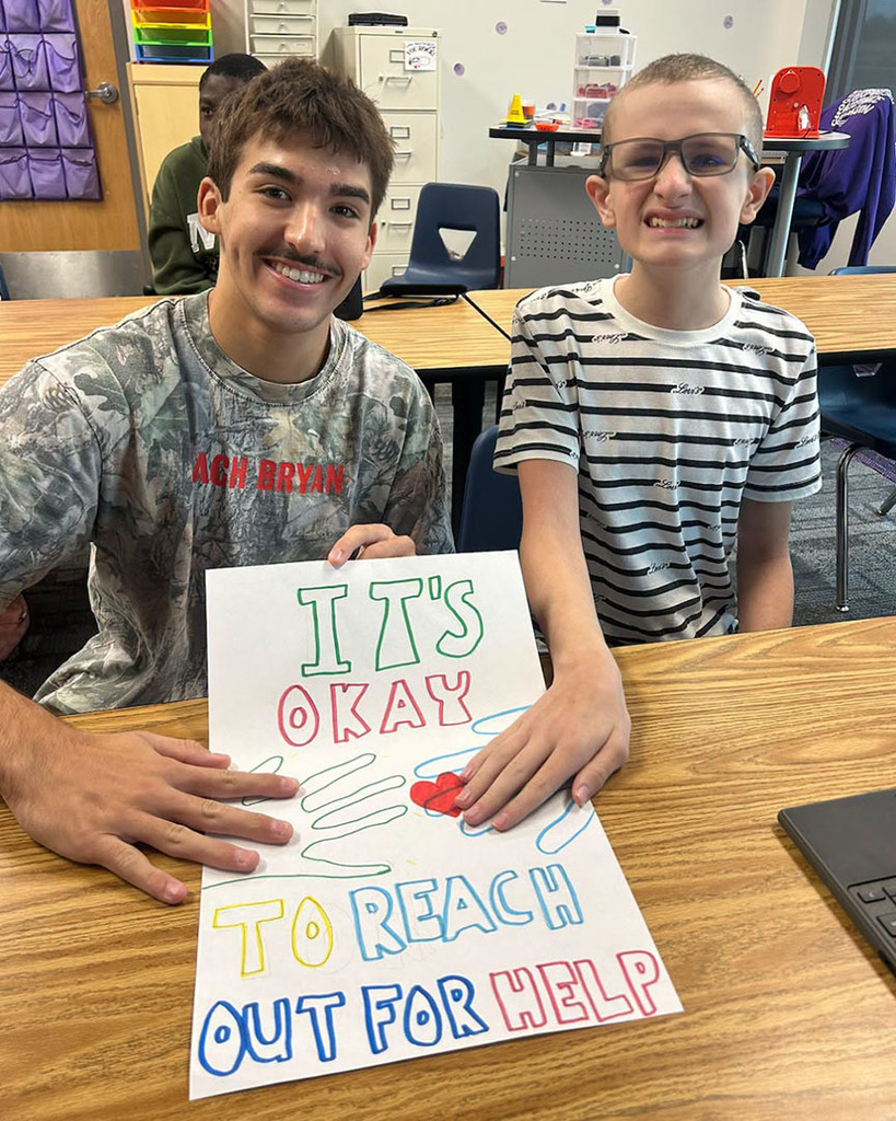 Two Northwest High School student are sitting at a desk in a classroom. They are holding a sign that says, "It's okay to reach out for help."