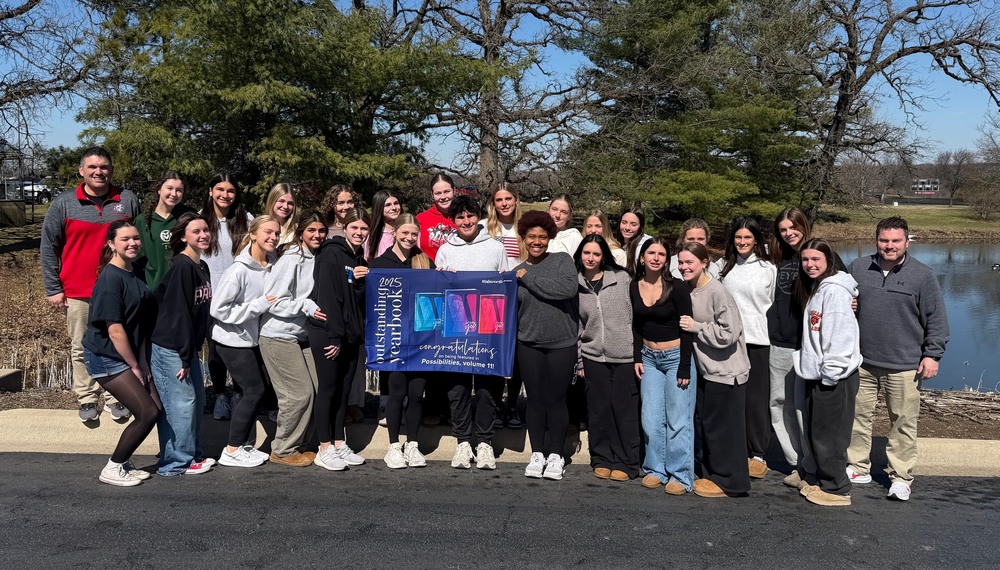 Students standing holding a banner in front of a pond. The banner recognizes their achievement in excellence in yearbook publishing.