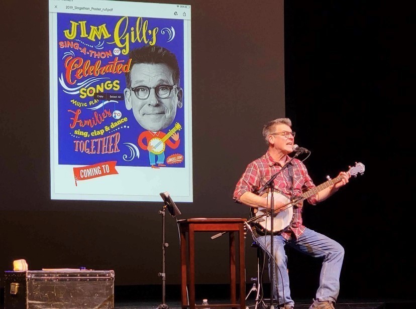 Jim Gill sitting on stage signing a song with a guitar