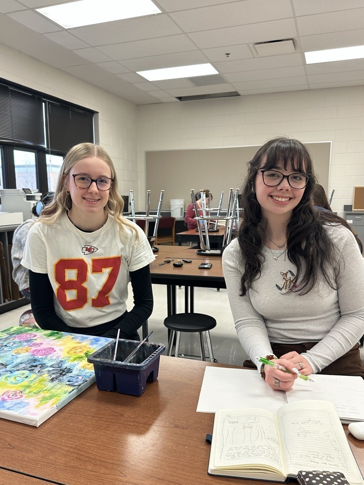 Two students sitting at a table in a classroom  with paintings and art supplies on it