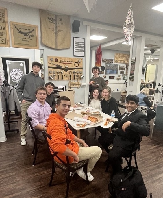 A group photo of students sitting and standing around a table eating pizza and drinking coffee