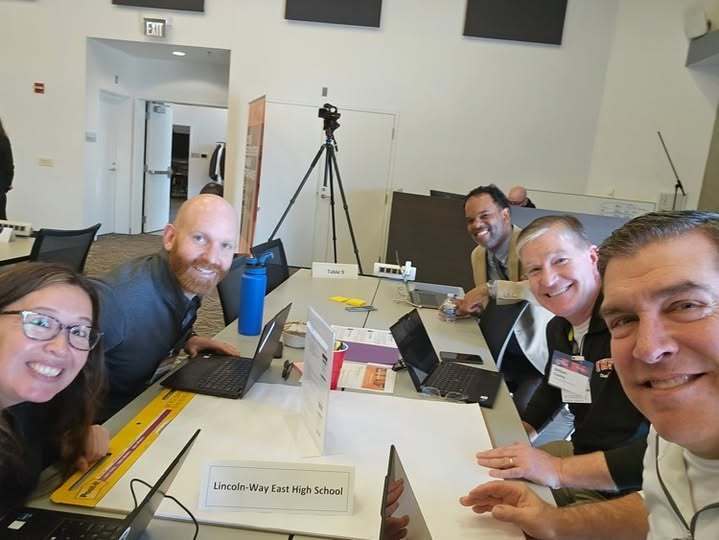 Teachers sitting around a table with laptops and papers on it, smiling for a photo. There is a sign that reads Lincoln-Way East High School on the table.