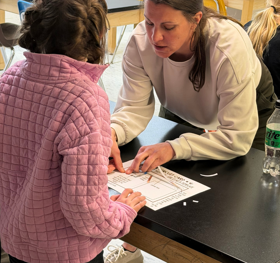 A woman helps a child with a craft project, both seated at a table. Background contains chairs and a table.