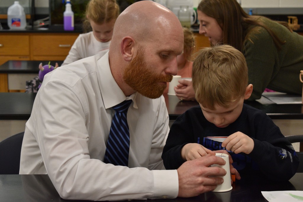 Man in a white shirt helping child in a black shirt do a craft. The child is holding a paper cup.