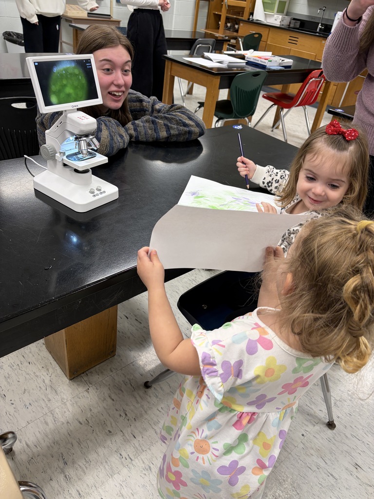 Two children stand by a microscope, one holding a paper, and another smiling. A table, chairs, and other equipment are in the background.