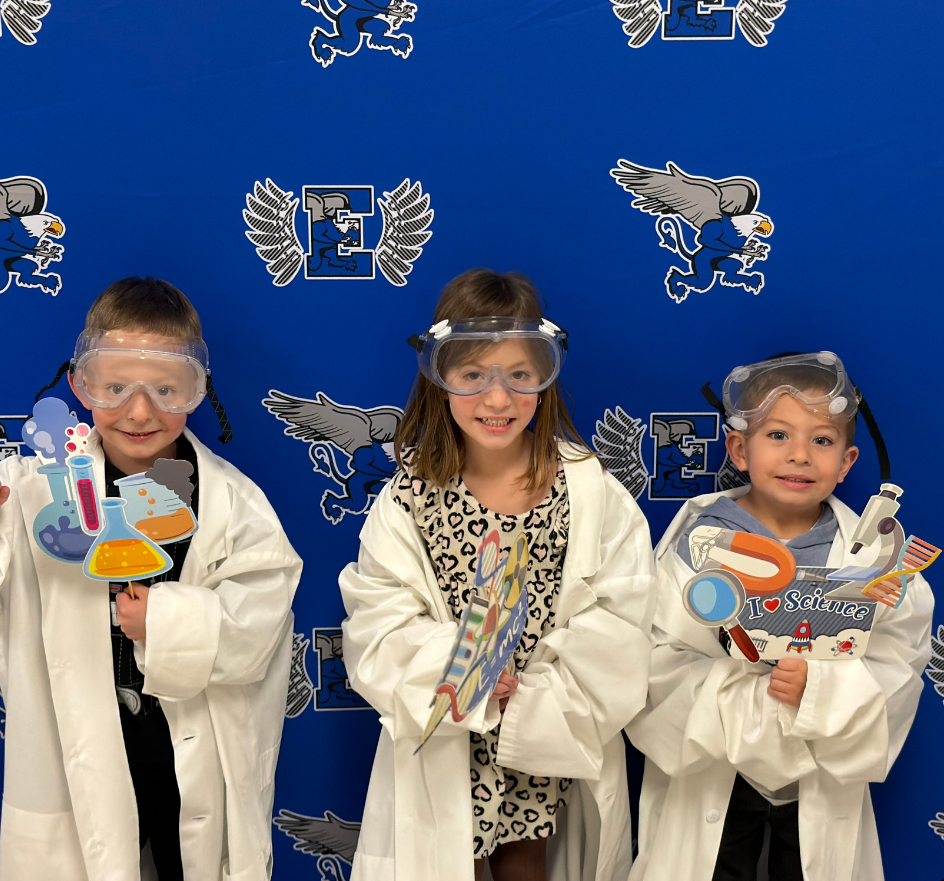 Three kids stand in front of a blue backdrop. They are wearing white lab coats and goggles.