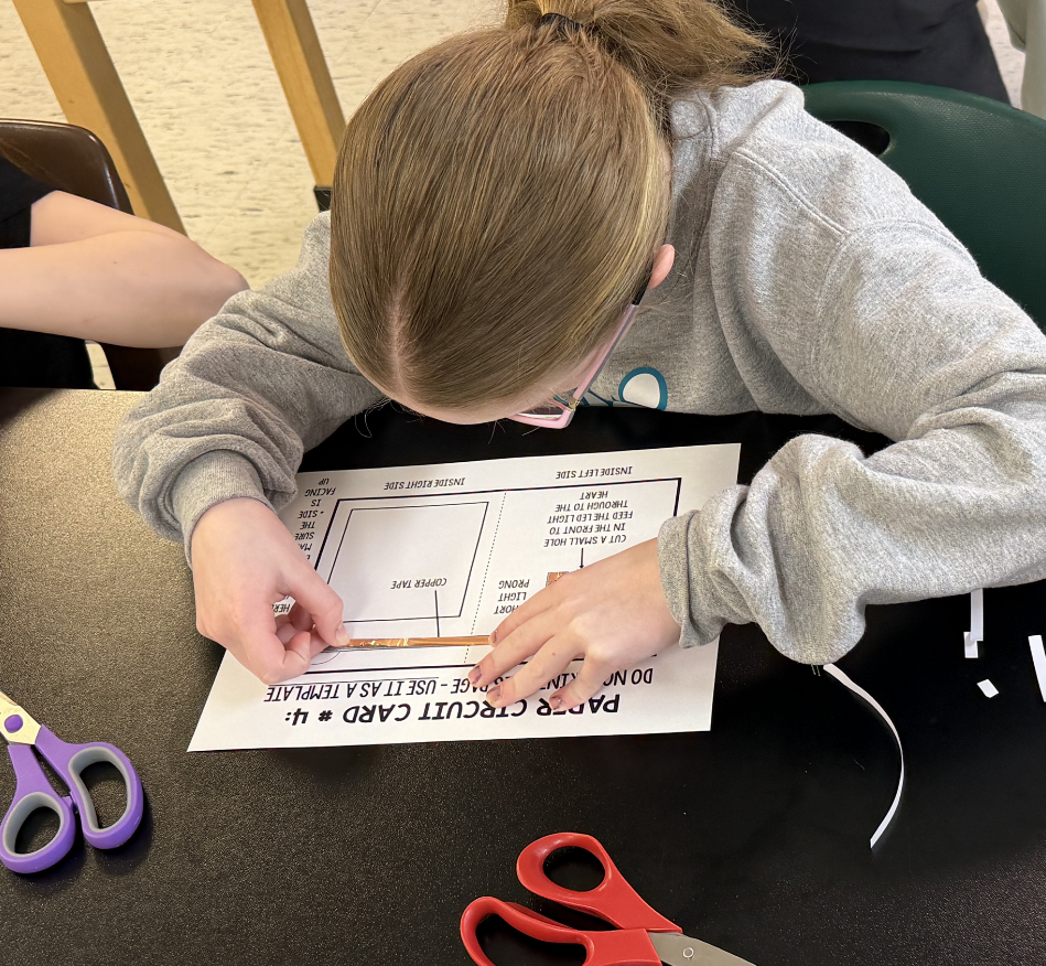 A young person in a grey sweater bends over a desk, handling scissors and a worksheet.