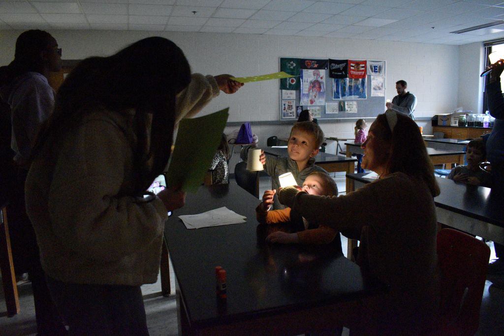 In a classroom, two students hold candles, while another holds a stick of yellow chalk. Background contains posters on walls and a desk.