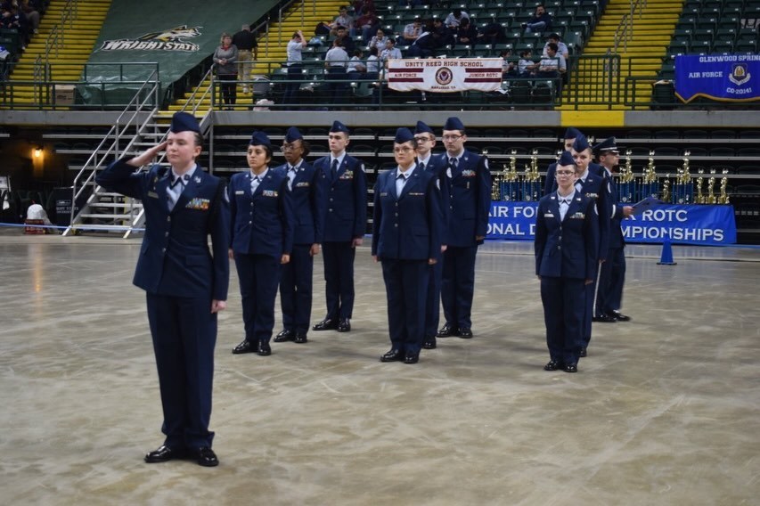 A group of uniformed individuals stands in a formation in an indoor arena. The individual in front salutes.