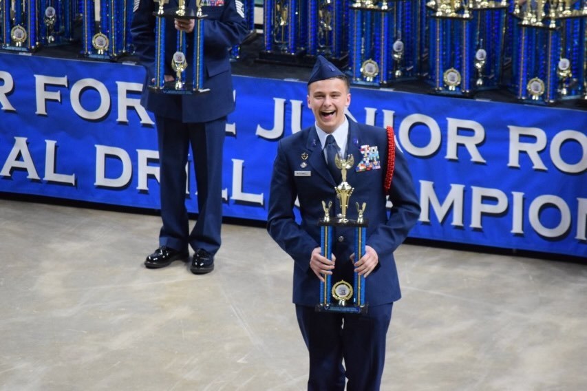 A student in a blue uniform holds a trophy while smiling in front of a banner.