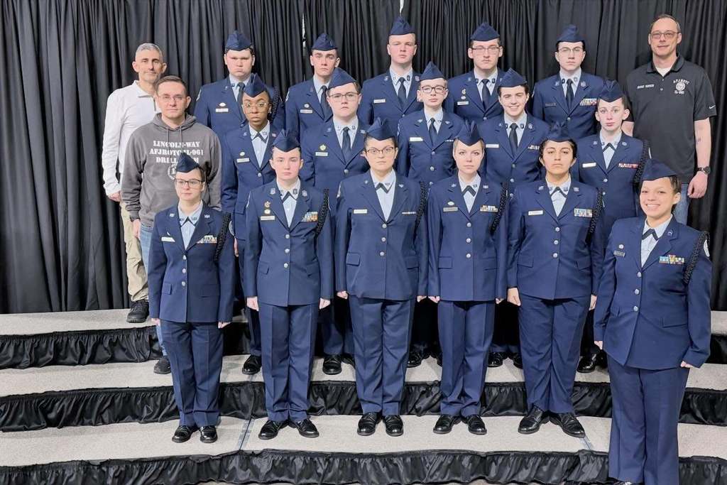 Group of uniformed individuals standing on a stage, some wearing hats, against a black curtain background.