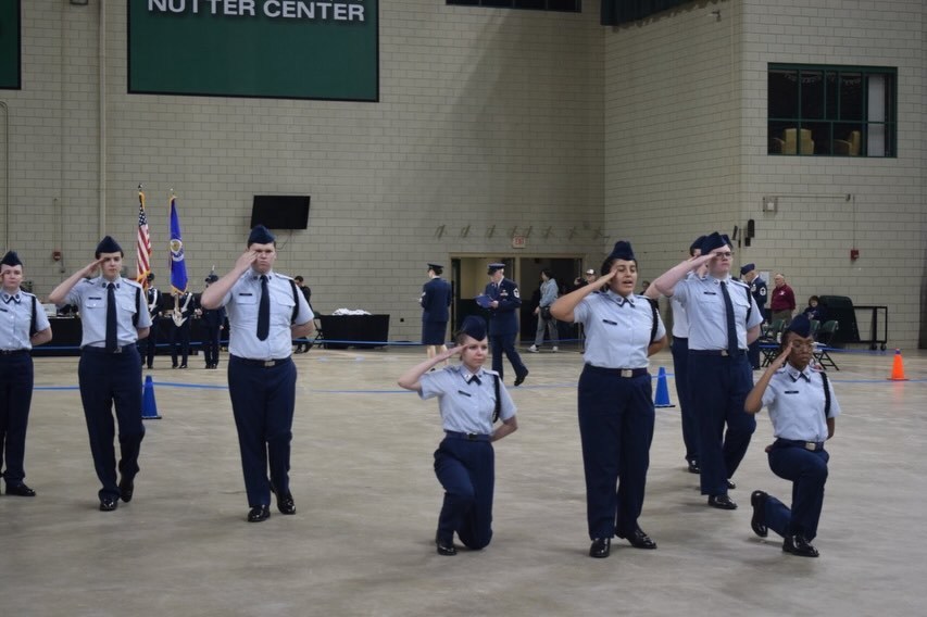 Military cadets perform a salute in a gymnasium with white walls and a green banner reading "NUTT CENTER".