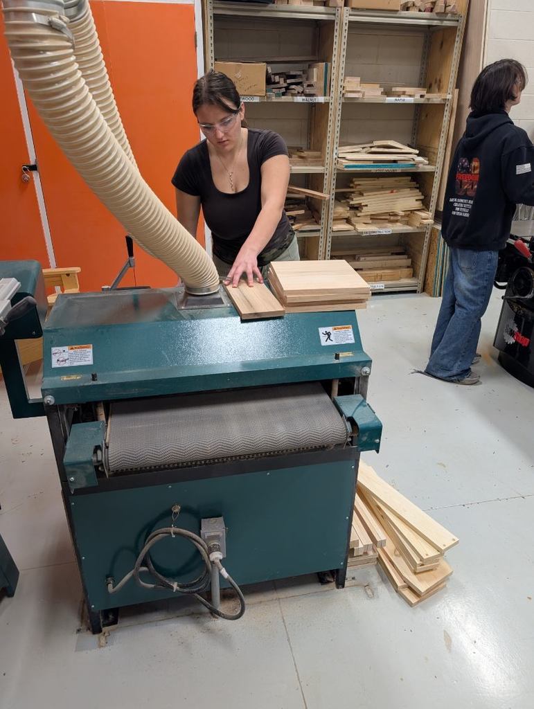 Two people in a workshop, one using a large machine to cut wood, another standing nearby.