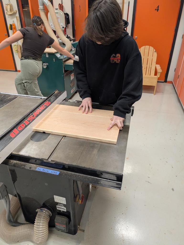 Two people in a workshop. One person bends over a table saw. Another person holds a wooden board.