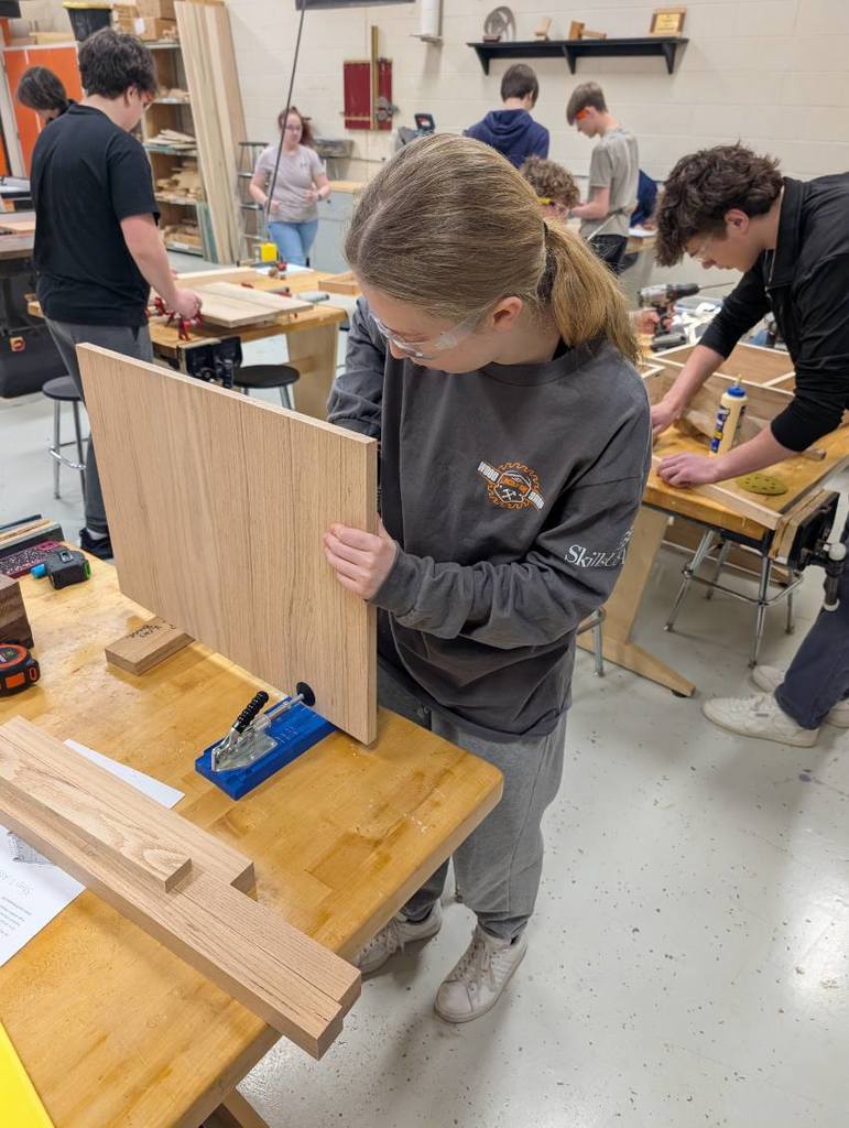 A group of people in a workshop. One person holds a wooden plank. Other people are working at tables.