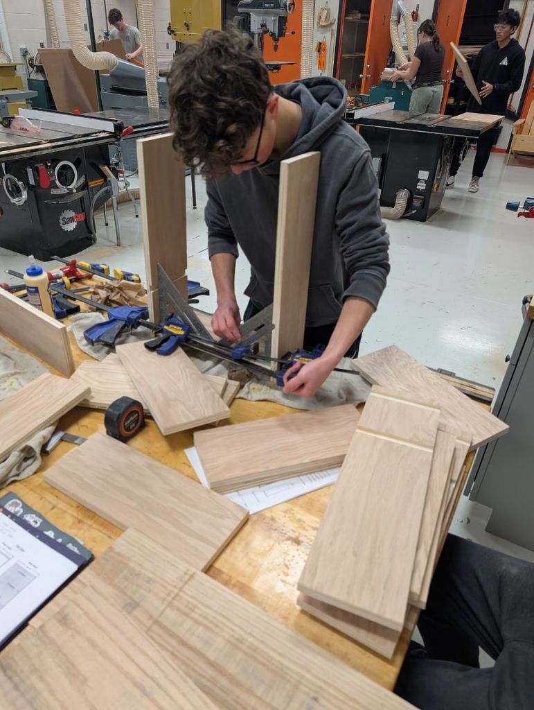 A person in a gray hoodie and glasses works on a wooden project at a workbench.