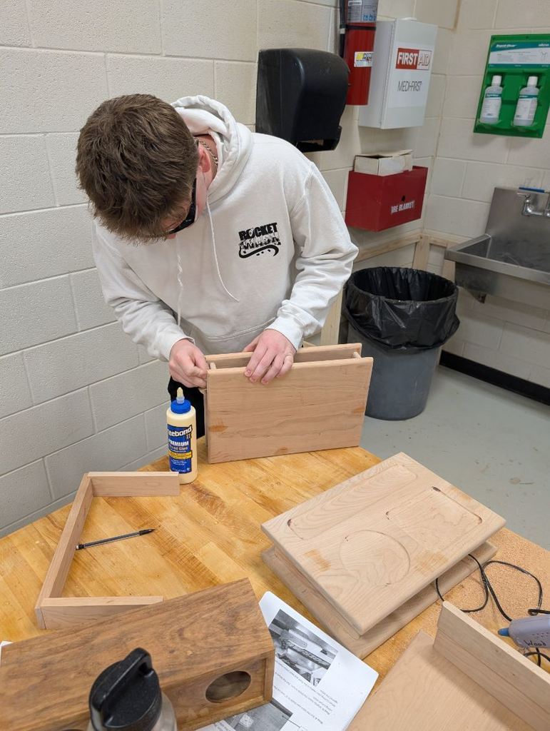 A person in a white hoodie working on a wooden box on a table. The background has a sink, trash bin, and dispensers.