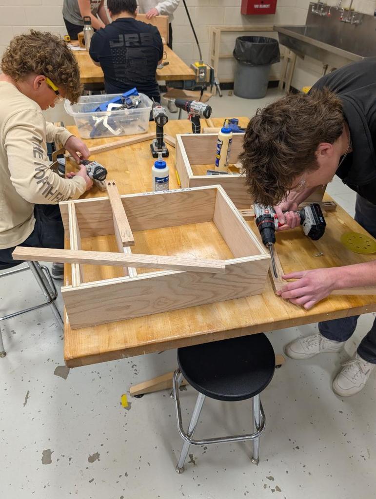 A group of people are working on wooden boxes in a room. One person uses a power drill on a wooden board.