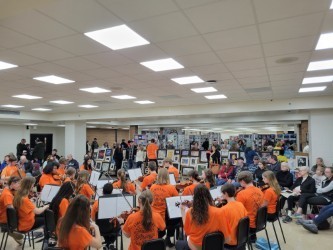 A group of people in orange shirts sit in chairs around a room and play instruments.