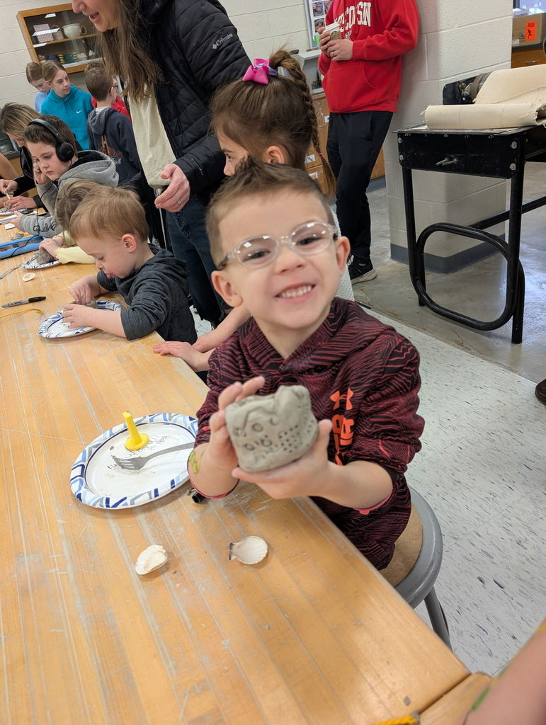 A child sits at a table holding a clay item, with other children seated behind them.