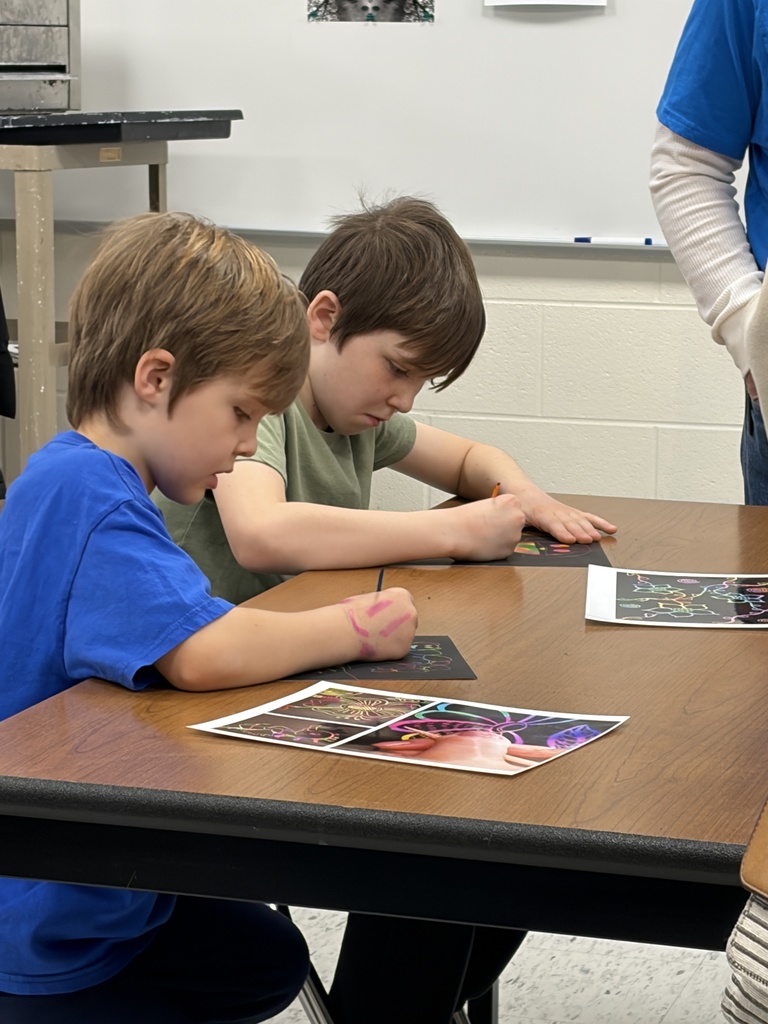 Two boys seated at a table, coloring pictures with pencils. One boy wears a blue shirt.