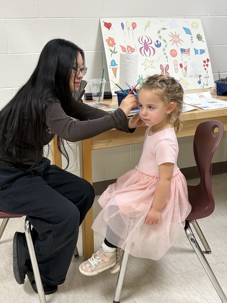 A woman helps a young girl with her face paint sitting on a chair next to a table.