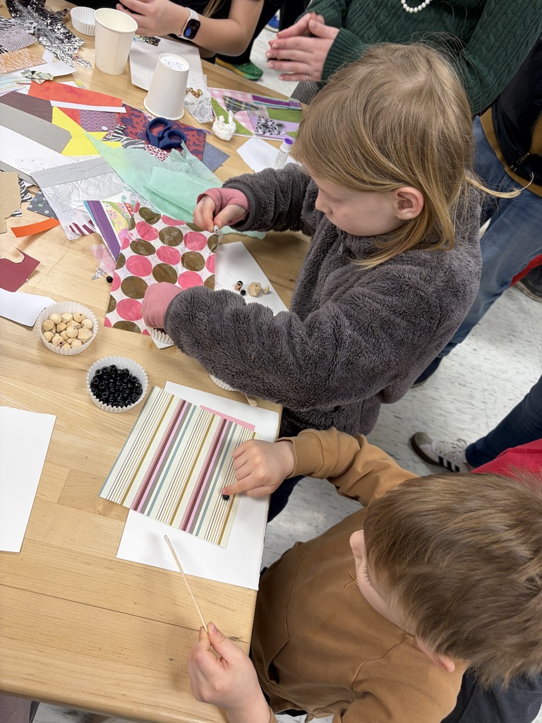 Children work on a craft project at a table, using paper, scissors, and beads. A girl is holding a paper with a polka dot design.