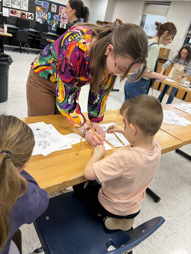 A woman and a boy are seated at a table, focusing on a craft project. A girl sits behind them, observing. Background: A classroom with tables, chairs, and walls adorned with artwork.
