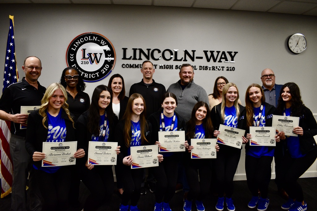 The Board poses with the East competitive cheer team, who are holding certificates and standing in front of a white wall with a flag, clock, and sign reading "LINCOLN-WAY".