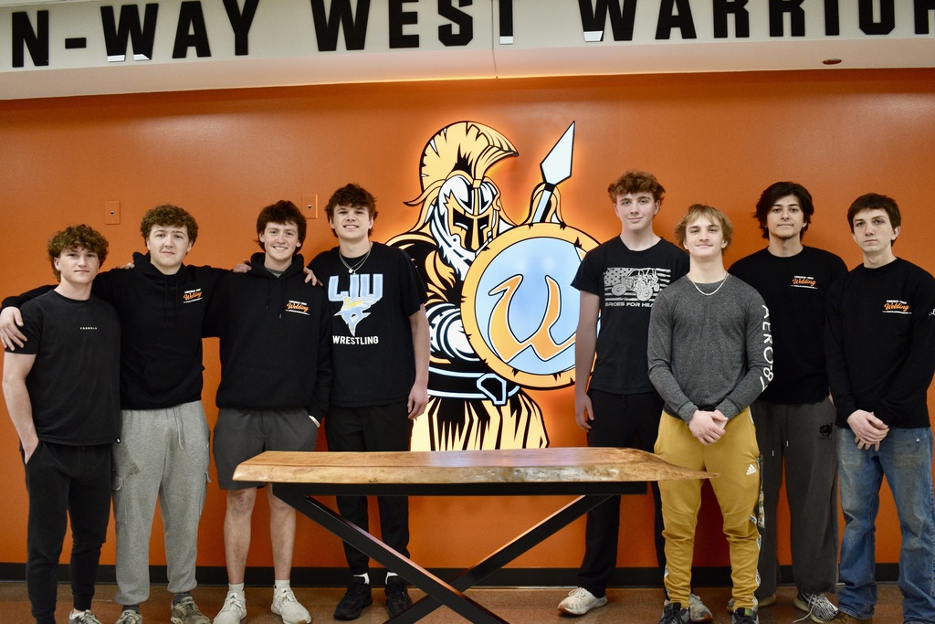 Nine boys in black t-shirts and jeans stand around a wooden table, with a wall behind them that says "Lincoln-Way West Warriors". In front of them is the live-edge table they created.