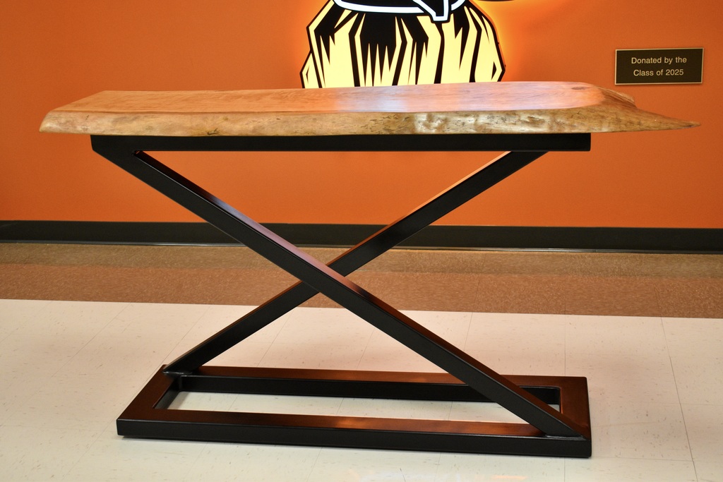 A modern wooden table with an angular, X-shaped metal base sits against an orange wall with a large orange warrior and a plaque.