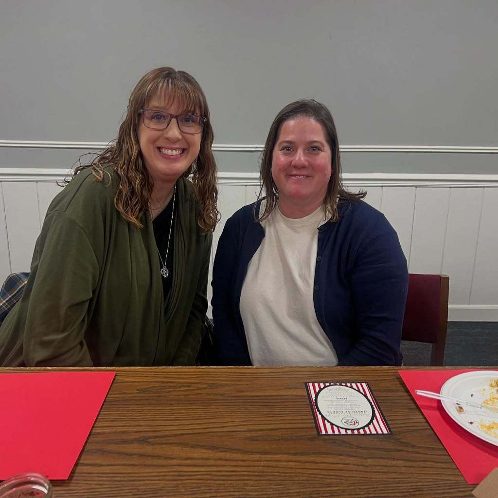 Two women sit at a table, smiling. Behind them is a wall with white trim.