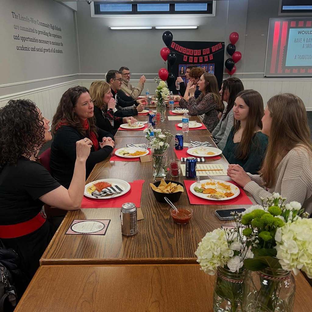 A group of people seated around a long table, enjoying food and drinks, with balloons and a screen in the background.