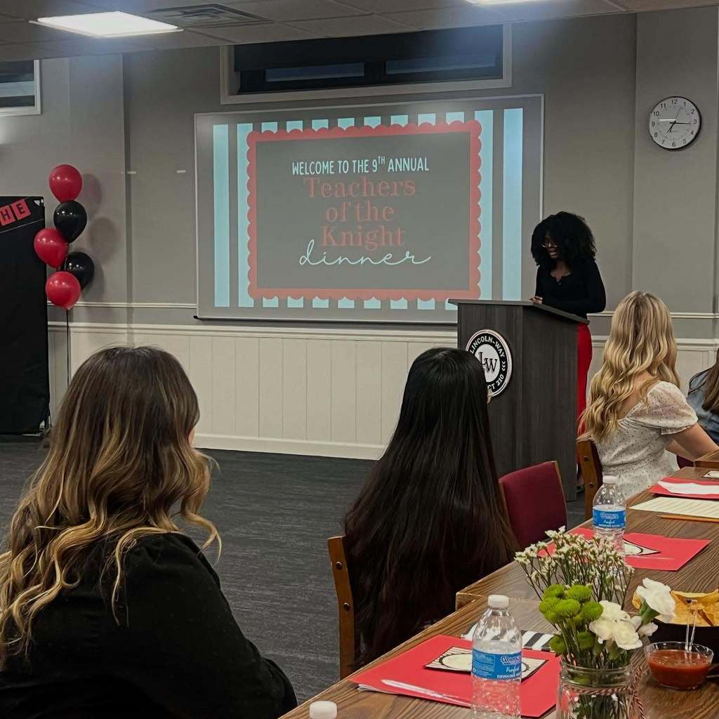 People sit in a classroom. A projector screen shows text. A student stands behind a podium.