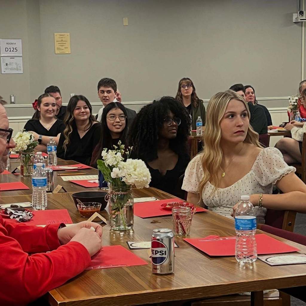 Group of people sitting around a table with red placemats, a can, and bottles of water.