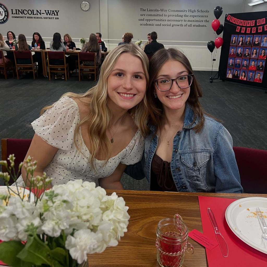 Two students sit at a table with a flower arrangement, smiling and wearing glasses. Behind them, a group of people sit at tables at an event.