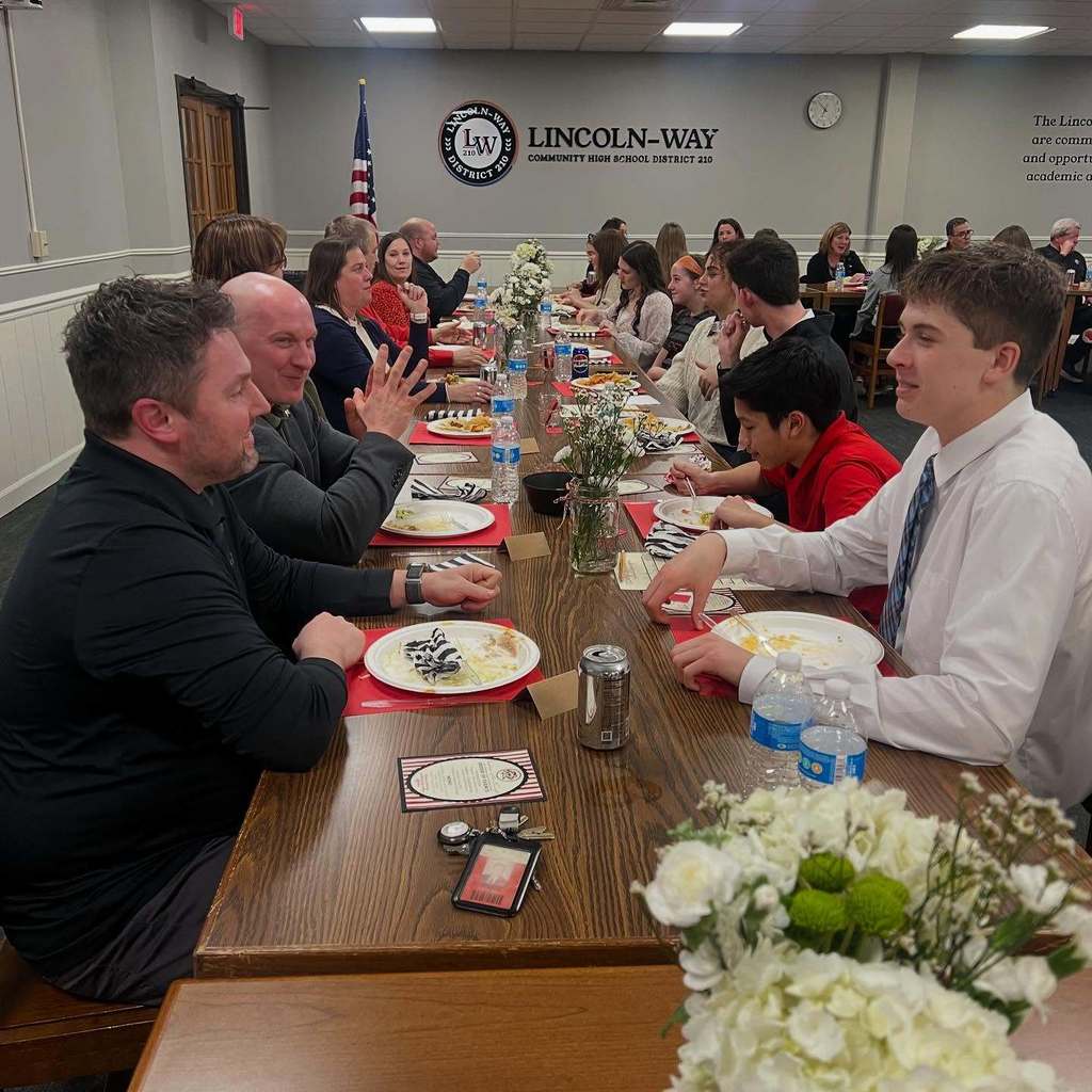 People seated at a long table in a meeting room, with plates, flowers, and cups.