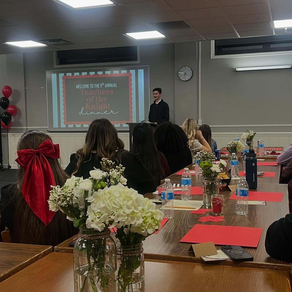 A group of people in a room with a projector screen. A student stands by a table with flowers.