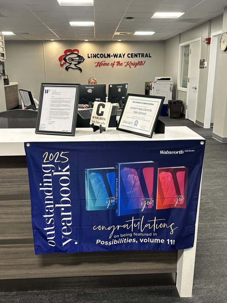 A reception desk displays a blue banner celebrating the 2015 yearbook. Framed certificates are on the desk.