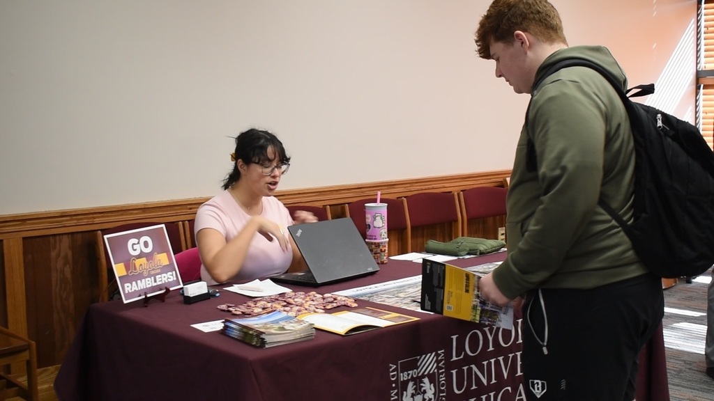 A person with a backpack stands by a table with a laptop. Another person sits, wearing glasses.