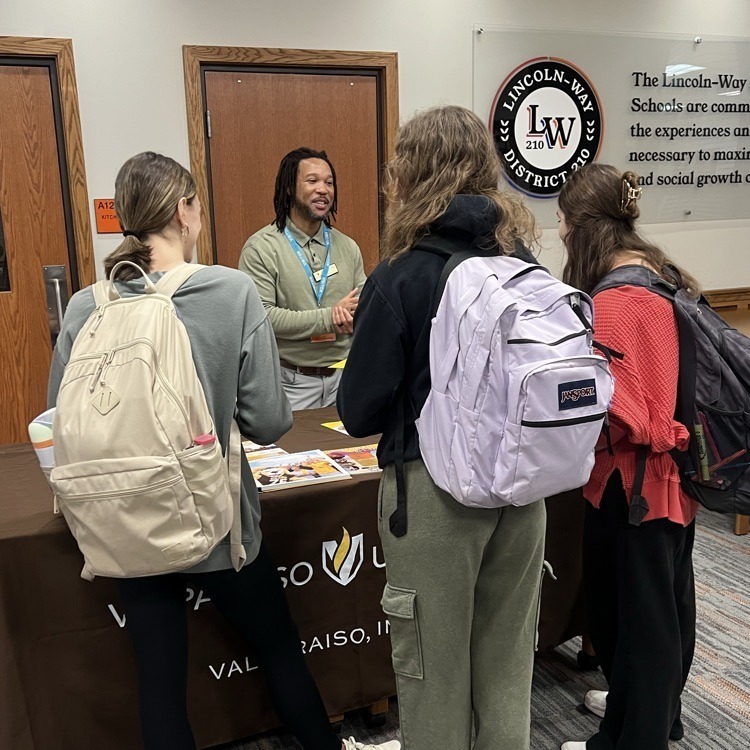 Four students wearing backpacks stand at a table, engaging with a man in a white shirt.