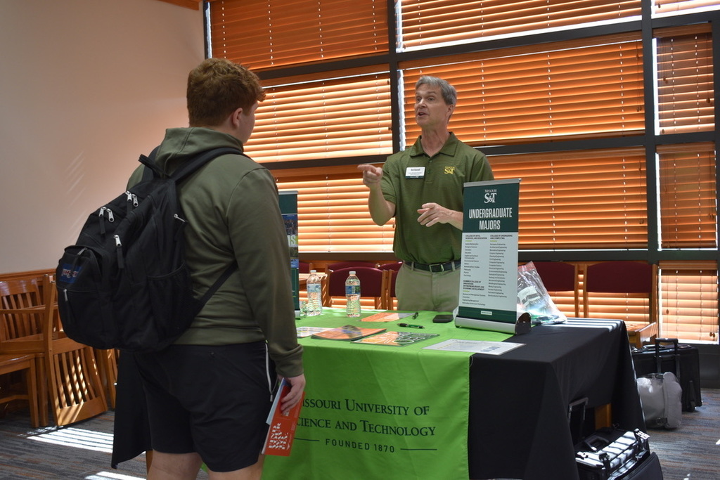 Two individuals engage in conversation at an event; one wears a green shirt, and the other wears a backpack.