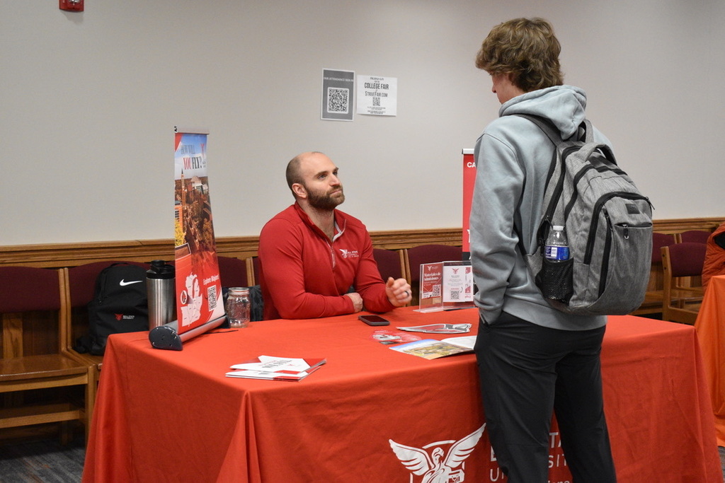 A man in a red shirt sits at a table, facing another man in a gray hoodie with a backpack.